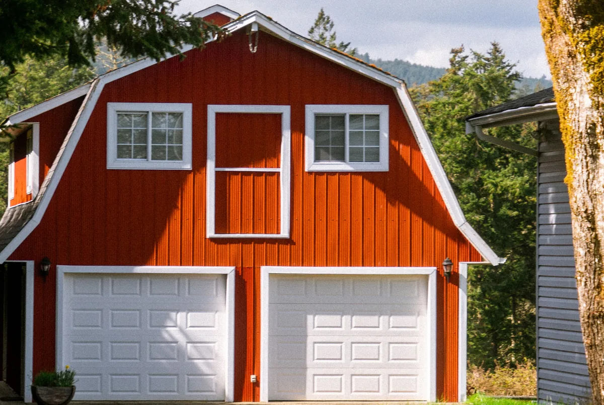 A red gambrel-roof barn with white garage doors and trim beside a wooded green hillside