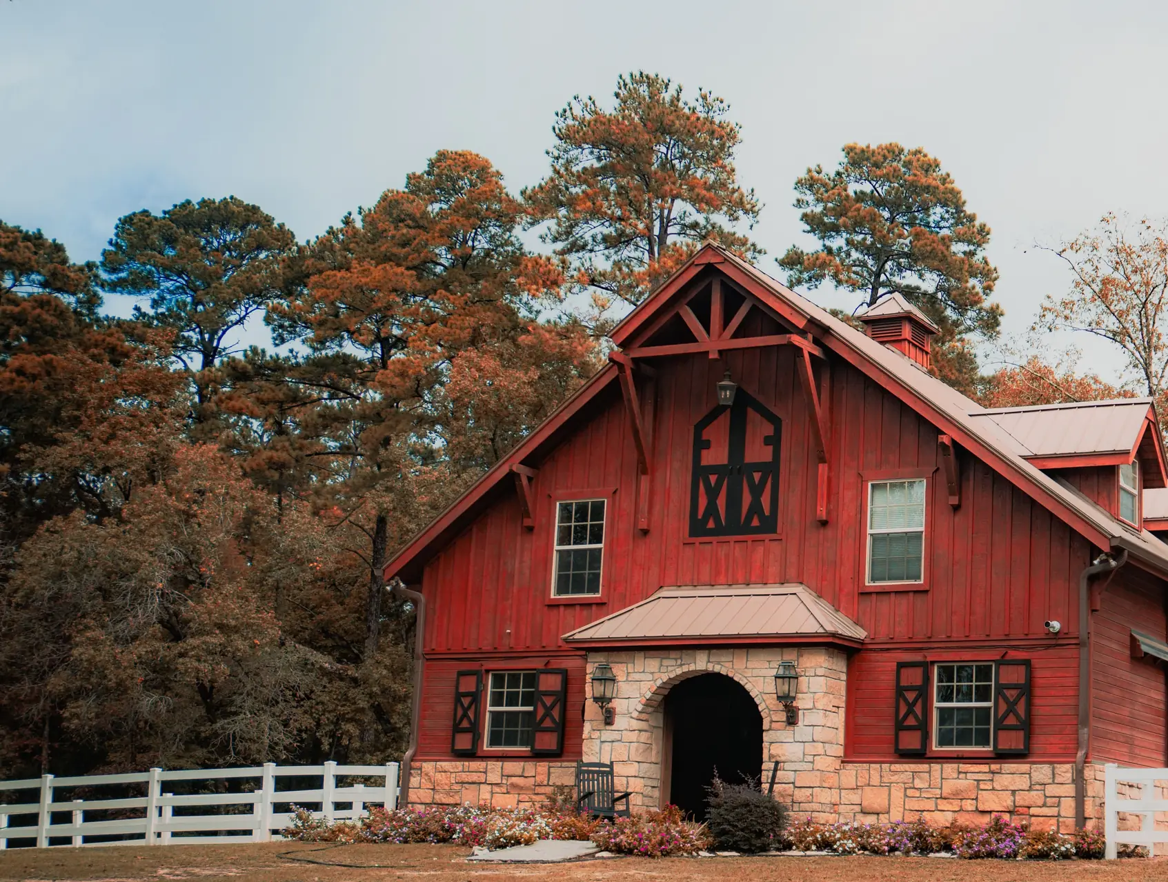 A red timber-frame barn-style home with a gabled metal roof, stone entry arch, and a white rail fence in a wooded Tennessee clearing